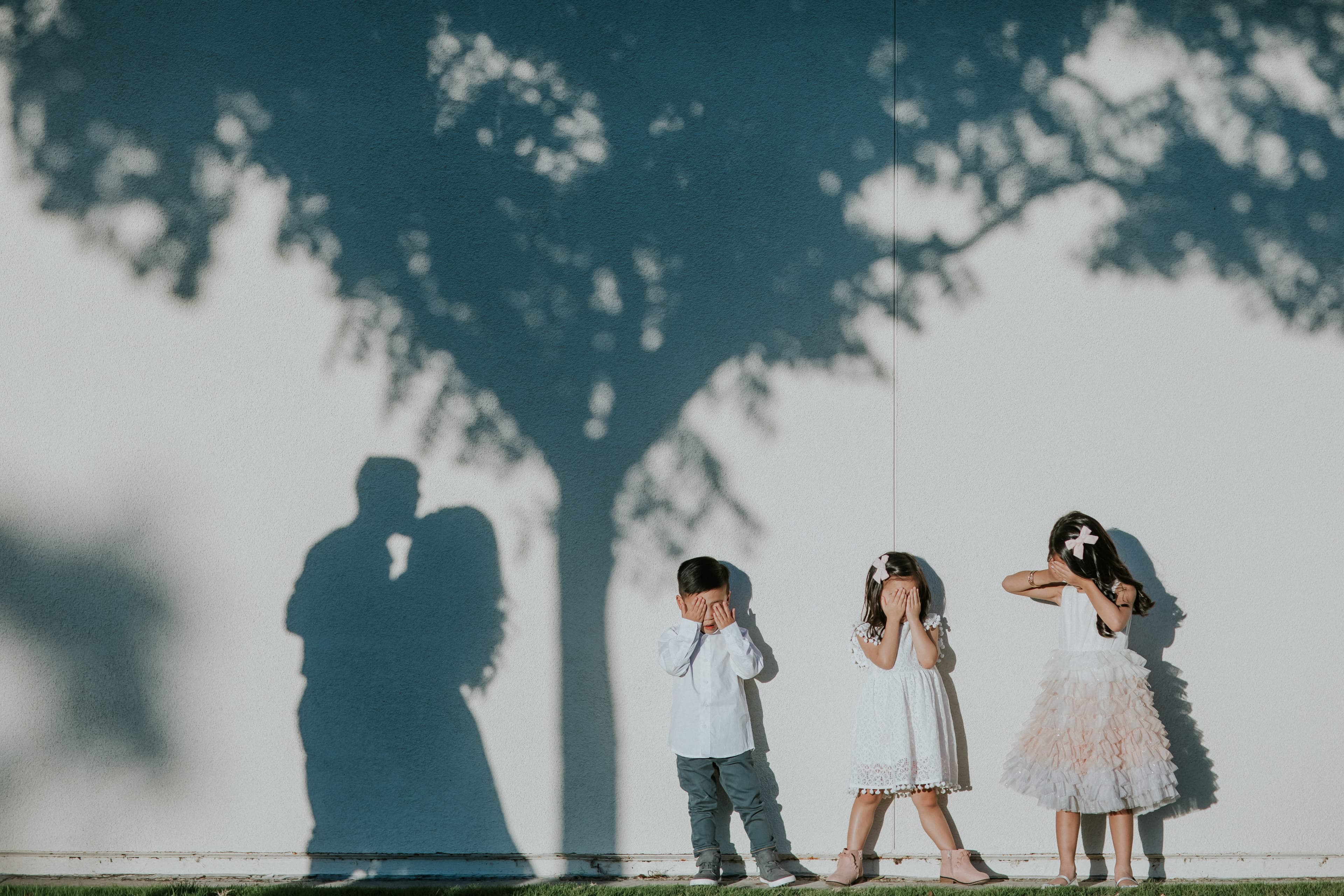 Three children cover their eyes against a white wall; the shadows of their parents kiss beneath a tree.